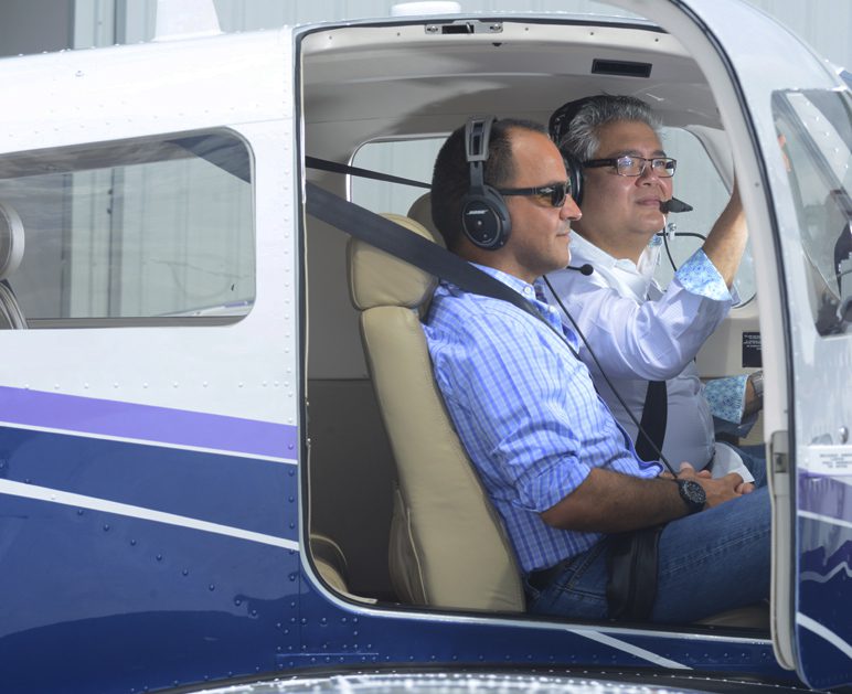 Two men seated in the cockpit of a small aircraft, each wearing headsets. The man on the right is holding the control yoke and the man on the left is observing.