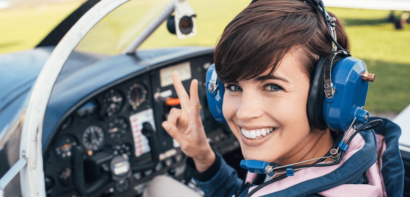 A woman wearing a headset and smiling shows a peace sign while sitting in the cockpit of an aircraft.