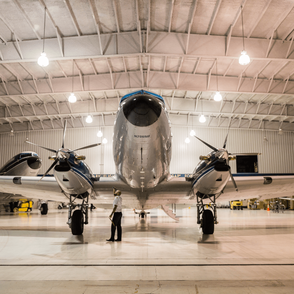 A person stands near the nose of a twin-engine aircraft in a hangar, with the plane illuminated by overhead lights.