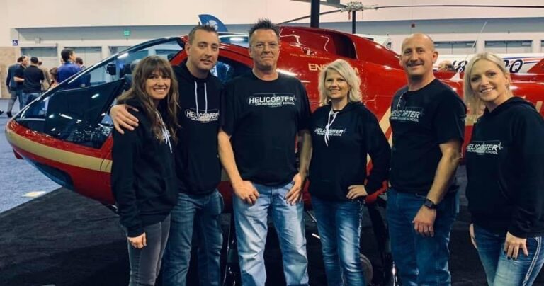 A group of five people wearing black hoodies with "HELICOPTER" text stands in front of a red helicopter inside an exhibition hall.