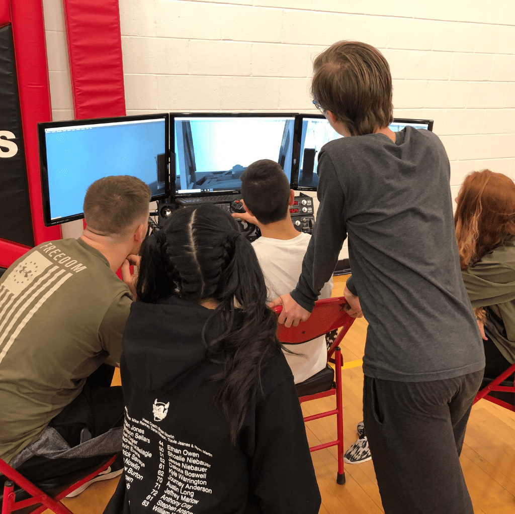 A group of young people gathers around a computer setup with multiple monitors in a gym. Two of them are seated, actively using the controls, while the others watch.