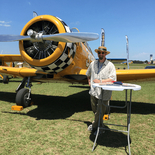 A man stands next to a yellow, vintage aircraft on display outdoors. He is wearing sunglasses, a hat, and a patterned shirt, with a folding table in front of him. The sky is clear and blue.