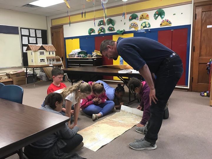 A man leans over to discuss a large map spread out on the floor with a group of six children in a classroom setting.