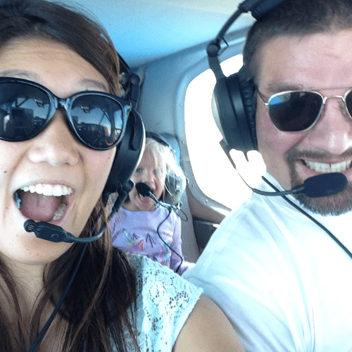 Three people wearing sunglasses and headsets, appearing excited, are seated inside a small aircraft. One person is a child positioned in the back.