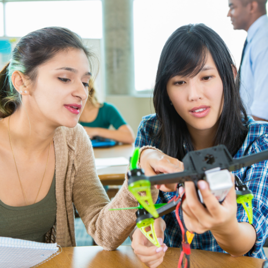 Two women in a classroom work together on assembling a drone, with one pointing and explaining, while a man in the background observes.