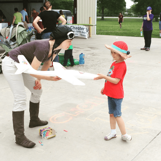 An adult dressed as an aviator hands a child in a red shirt and hat a model airplane during an outdoor event. A chalk drawing with "Check in Here" is visible on the ground.