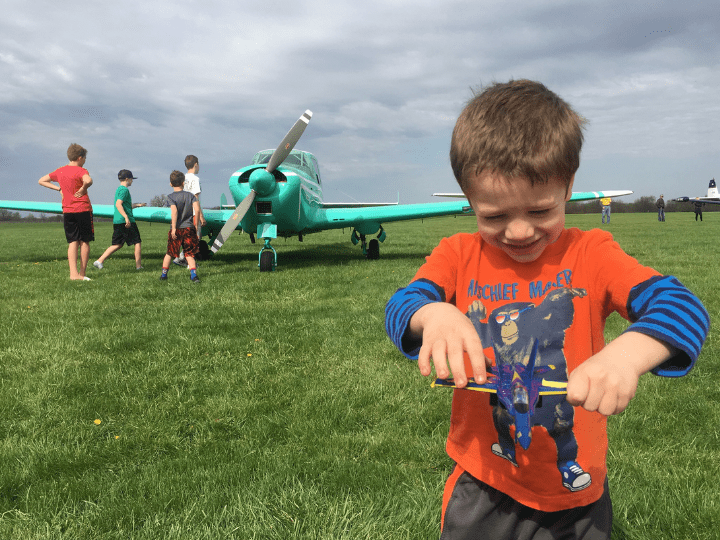 A young boy in an orange shirt plays with a toy plane on grassy field while several other children gather near a small green aircraft in the background under a cloudy sky.