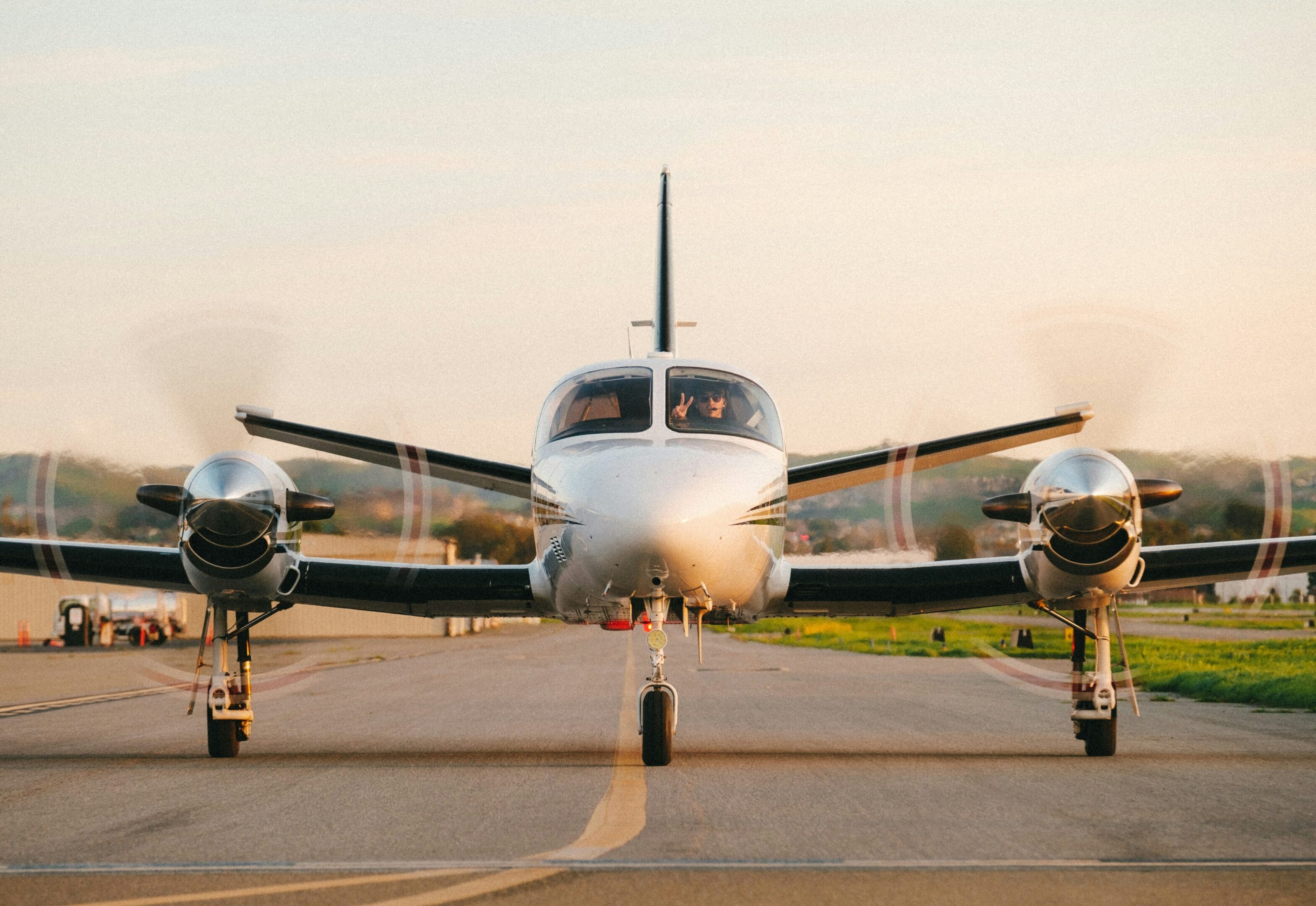 An Open Air Flight Club Plus, a compact twin-engine airplane with spinning propellers, is stationary on a runway, facing directly forward on a clear day.